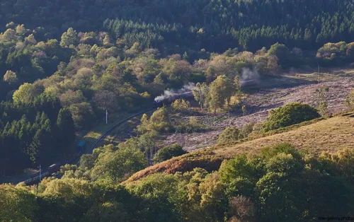 Autumn steam This landscape photograph captures the scenic rural environment of the North Yorkshire Moors in the United Kingdom during autumn 2018. Taken in the afternoon, the image prominently features a steam train moving through the moorland, surrounded by nature and a patchwork of mixed woodland and open areas. Wisps of steam mark the train’s progress along the curved track, highlighting the theme of autumn steam transport against the backdrop of changing foliage. The landscape is rich in autumn colours, with golden and russet tones evident in the trees and undergrowth, illustrating the transition of the season in this rural setting. This photograph showcases the harmony between historical transport and the natural environment in North Yorkshire Moors, emphasising the beauty of the autumnal countryside in 2018.